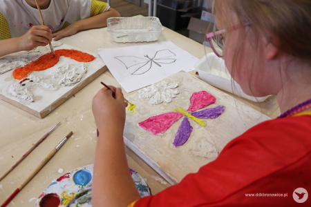 Kolorowe zdjęcie. Zbliżenie na stół. Na stole leżą prace wykonane przez dzieci. Dłoń dziewczynki maluje pędzlem płaskorzeźbę motyla.