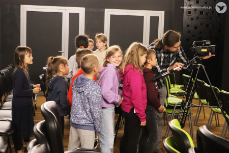 Kolorowe zdjęcie. Grupa dzieci stoi między krzesłami zaglądając w kamerę. Przy kamerze stoi instruktor DDK Bronowice.