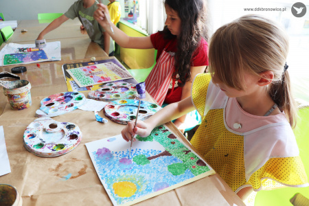 Kolorowe zdjęcie. Dziewczynki siedzą przy stoliku. Malują farbami impresjonistyczne obrazy. Na stole leżą paletki z farbami.