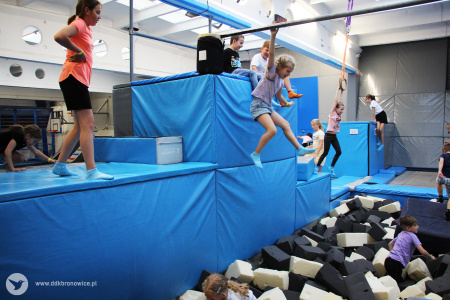 Kolorowe zdjęcie. Park trampolin. Pomieszczenie z materacowymi, niebieskimi klockami i basenem czarno-białych pianek. Dzieci skaczą z klocków do basenu.