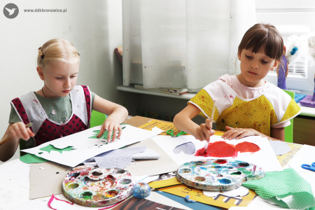 Kolorowe zdjęcie. Dwie dziewczynki siedzą przy stole. Malują farbami na wyciętych z materiału chmurach z pomocą szablonów. Na stole leżą palety z farbami.