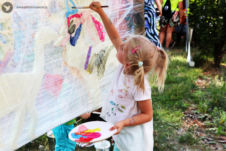 Kolorowe zdjęcie. Plener. Dziewczynka trzyma w lewej dłoni talerzyk z farbami.  W prawej dłoni trzyma pędzel i maluje na rozciągniętym pomiędzy drzewami stretchu motyla ze styroduru.
