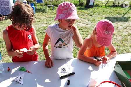 Kolorowe zdjęcie. Chłopiec i dwie dziewczynki stoją przy stoliku warsztatowym i wykonują origami.