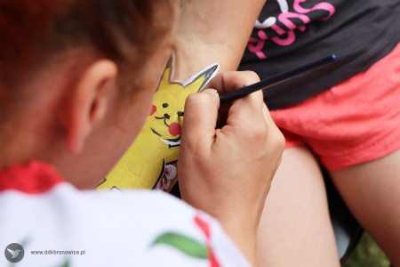 Kolorowe zdjęcie. Kobieca dłoń maluje pędzlem tatuaż na przedramieniu. Malowana postać to pikachu.