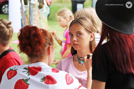 Kolorowe zdjęcie. Kobieta maluje farbami wzór na szyi dziewczyny. Dziewczyna kieruje wzrok na fotografa.