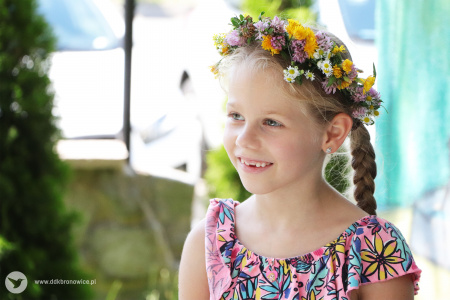 Kolorowe zdjęcie. Dziewczynka w wianku z kwiatów na głowie. Patrzy się w bok i uśmiecha się.