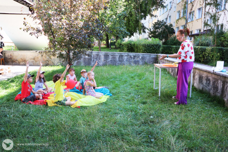 Kolorowe zdjęcie. Podwórko, zielona trawa. Po prawej stronie instruktorka DDK Bronowice stoi bokiem do fotografa i tłumaczy coś dzieciom. Naprzeciwko niej na kolorowej chuście siedzą dzieci i podnoszą rękę do góry.