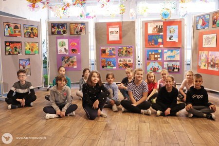 Kolorowe zdjęcie. Uczniowie ze szkoły podstawowej nr 31 w Lublinie siedzą na podłodzie po turecku. Uśmiechnięci pozują do zdjęcia. W tle stoją tablice z ich pracami plastycznymi.