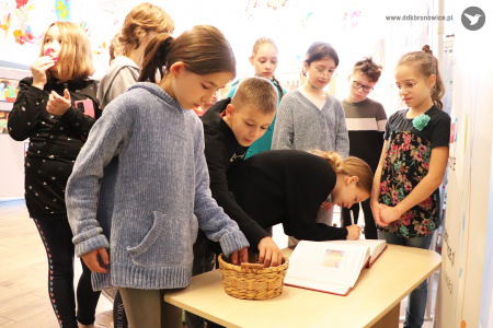 Kolorowe zdjęcie.  Dzieci stoją przy stoliku z pamiątkową księgą. Dziewczynka z chłopcem sięgają dłońmi do koszyczka z cukierkami. Jedna dziewczynka podpisuje się w księdze.