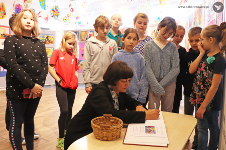 Kolorowe zdjęcie. Nauczycielka ze szkoły podstawowej nr 31 w Lublinie wpisuje się siedząc przy stole do pamiątkowej księgi Domu Kultury. Dzieci gromadzą się wokół niej i patrzą w kierunku księgi.