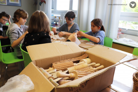Kolorowe zdjęcie. Z przodu w kartonowym pudle drewniane wałki. W tle przy stolikach  siedzą dzieci  i wykonują czynności plastyczne.