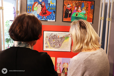 Kolorowe zdjęcie. Zdjęcie zrobione od tyłu. Dwie kobiety oglądają prace plastyczne wywieszone na tablicy.