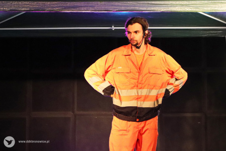 Kolorowe zdjęcie. Aktor w pomarańczowym kombinezonie stoi na scenie z podpartymi na boki rękoma.