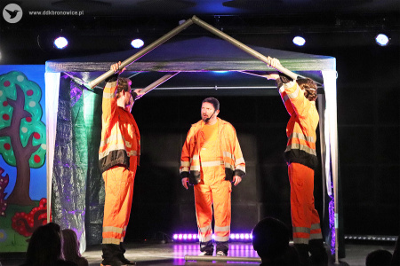 Kolorowe zdjęcie. Trzech aktorów w pomarańczowych kombinezonach. Dwóch z nich stojących po bokach trzymają wysoko w górze w dłoniach rury tworząc dach. Aktor stojący pomiędzy nimi przemawia, a dłonie ma spuszczone wzdłuż ciała.