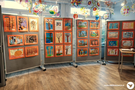 Zdjęcie kolorowe. Na czterech tablicach wystawienniczych wiszą prace dzieci o tematyce jesiennej. Po prawej stronie na stole leży księga pamiątkowa. Z sufitu zwisają kolorowe ozdoby.