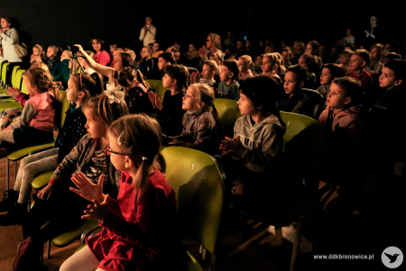 Zdjęcie kolorowe. Liczna publiczność złożona z dzieci bije brawo siedząc na krzesłach ustawionych w rzędach.