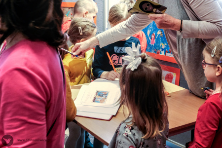 Zdjęcie kolorowe Dzieci stoją przy stole z księgą pamiątkową. Kobieta w wyciągniętej ręce trzyma długopis.