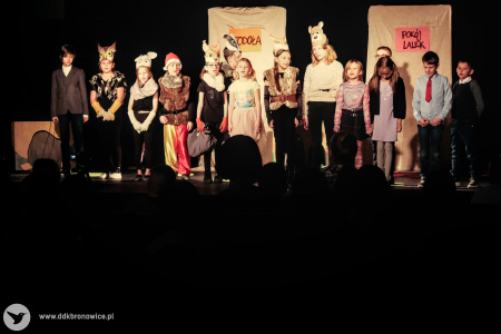 zdjęcie kolorowe. Na oświetlonej scenie przebrane dzieci stoją w rzędzie. mają otwarte usta, można się domyślić, że śpiewają lub mówią synchronicznie.