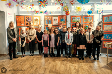 Zdjęcie kolorowe. Dzieci, nauczycielki i instruktorka DDK Bronowice stoją w dwóch rzędach w Galerii Małej na tle tablic wystawienniczych z pracami. Uśmiechają się do zdjęcia.