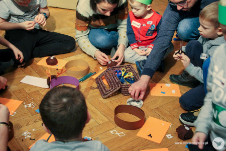 Kolorowe zdjęcie. Dzieci z rodzicami siedzą na podłodze. Przed nimi leżą nożyczki, opaski z papieru oraz pomarańczowe pianki. Tworzą opaski zwierzęce.