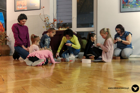 Kolorowe zdjęcie. Dwie mamy i piątka dzieci siedzą na podłodze. Obok nich siedzi animatorka. Wykonują opaski zwierzęce.