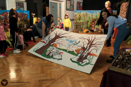 Kolorowe zdjęcie. Animatorki wnoszą na salę wielkoformatową planszę z namalowanymi elementami lasu. Wokół stoją dzieci w opaskach ze zwierzątkami. W tle dekoracja jesienna.