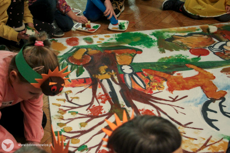 Kolorowe zdjęcie. Pomalowany fragment wielkoformatowej planszy. Widać namalowane drzewo, zająca, jelonka i grzyby. Wokół niej są dzieci.