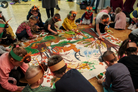 Kolorowe zdjęcie. Na podłodze leży wielkoformatowa plansza. Namalowane są na niej drzewa, jelonek, zając i grzyby. Dzieci zgromadzone wokół niej w opaskach zwierzęcych malują planszę.