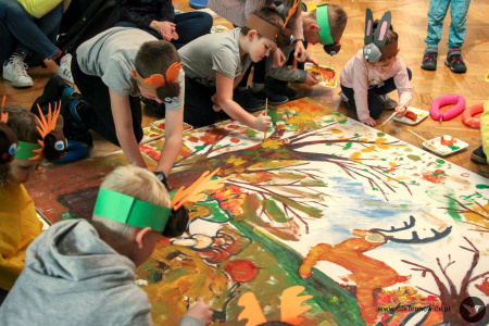 Kolorowe zdjęcie. Dzieci w opaskach zwierzęcych malują wielkoformatową planszę farbami.
