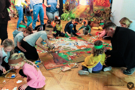 Kolorowe zdjęcie. Dzieci z w opaskach zwierzęcych wraz z rodzicami malują wielkoformatową planszę farbami. W tle widać dekoracje jesienne.
