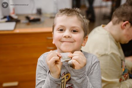 Kolorowe zdjęcie. Uśmiechnięty chłopiec pozuje do zdjęcia. W dłoniach trzyma mały skrawek papieru i pokazuje go.