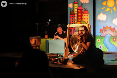 Zdjęcie kolorowe. Po prawej stronie na scenie siedzi Sylwetta, po lewej kuca mężczyzna. Przed nim stoi duży bęben. Obydwoje mówią lub śpiewają do mikrofonów. Sylwetta ma w ręku tłuczek do gongu.