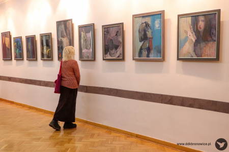 Zdjęcie. Na ścianie wisi szereg obrazów. Uczestniczka wystawy przygląda się jednemu z nich z bliska.