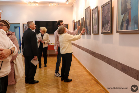 Zdjęcie. Uczestnicy wystawy spoglądają na obrazy. Kobieta z mężczyzną gestykulują i omawiają jeden z nich.