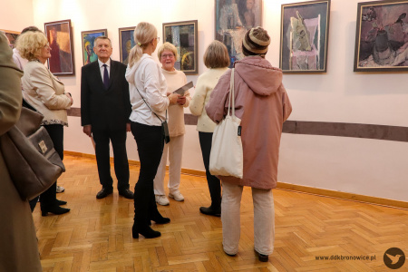 Zdjęcie. Uczestnicy wystawy wpatrują się w wiszące na ścianie obrazy. Artystka przemawia skierowana w ich kierunku.