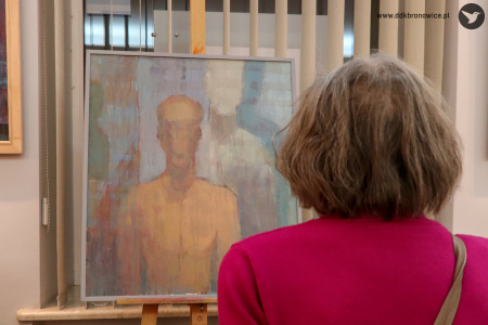 Zdjęcie. W centrum jeden z obrazów wystawy. Po prawej stronie sylwetka kobiety zwrócona tyłem wpatruje się w obraz.