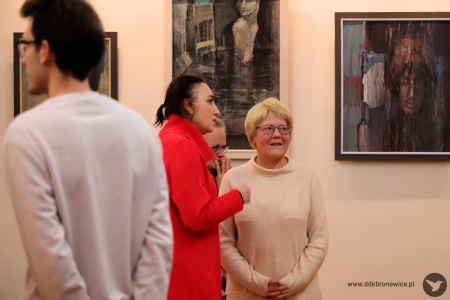 Zdjęcie. Artystka wraz z kobietą dyskutują wpatrzone w prawą stronę w kierunku obrazów znajdujących się na ścianie.
