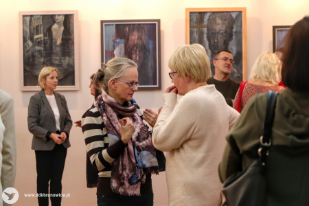 Zdjęcie. Artystka i kobieta rozmawiają stojąc bokiem. Artystka trzyma lewą dłoń przy ustach w geście zamyślenia. Kobieta wskazuje dłońmi na siebie. W tle stoją inni uczestnicy wystawy i na ścianie wiszą obrazy.