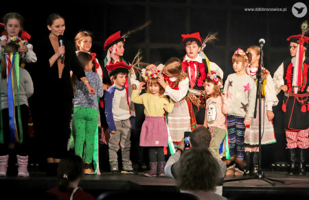 Zdjęcie kolorowe na scenie stoją dzieci w strojach ludowych, dziewczynki zakładają wianki dzieciom z publiczności. Kobieta z mikrofonem zwraca się do publiczności. Dorośli widzowie robią zdjęcia.