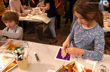 Zdjęcie kolorowe. Dziewczynka i chłopiec rysują i ozdabiają wzory na kolorowej folii. W tle inne stoły robocze i osoby przy nich.