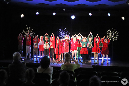 Zdjęcie. Grupa małych dzieci na scenie stoi w czerwonych ubraniach. Mają uniesione nad głową złączone dłonie.