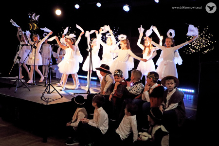 Zdjęcie. Dziewczynki w strojach aniołów tańczą z chustkami na scenie. Poniżej siedzą chłopcy w strojach pastuszków i króli.