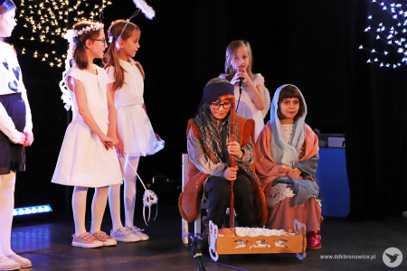 Zdjęcie. Dziewczynka w przebraniu Maryi oraz chłopiec w przebraniu Józefa siedzą nad kołyską z dzieciątkiem. Za nimi stoją dziewczynki przebrane za anioły. Jedna z nich recytuje do mikrofonu.