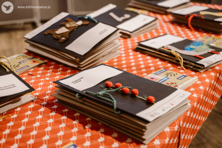 Zdjęcie kolorowe. Na stole, przykrytym czerwonym obrusem w biały wzór serduszek, leżą ułożone obok siebie książki sensoryczne różnej grubości i wielkości oraz foldery promocyjne projektu.