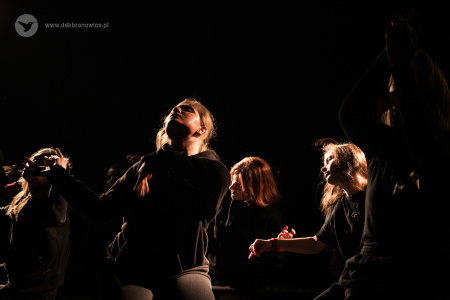 Kolorowe zdjęcie. Na zaciemnionej scenie grupa dziewczyn siedzi na scenie i patrzy w górę.