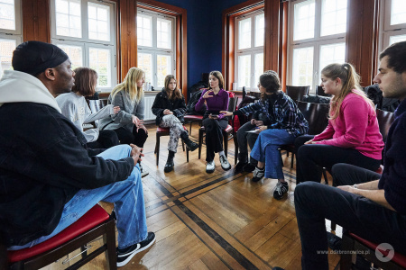 Kolorowe zdjęcie. Grupa młodych ludzi siedzi w półkole na krzesłach. Rozmawiają ze sobą.