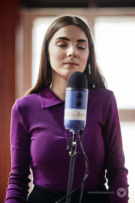 Kolorowe zdjęcie. Młoda kobieta stoi przed mikrofonem z zamkniętymi oczami.