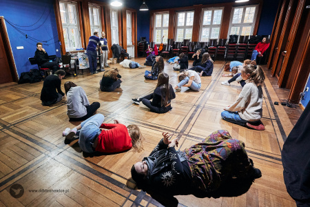 Kolorowe zdjęcie. Na drewnianej podłodze grupa młodych osób. Jedni siedzą po turecku, inni leżą na boku.