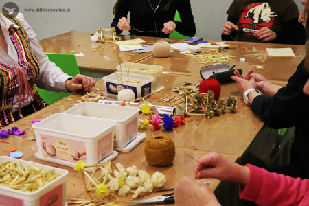Zdjęcie. Na stole warsztatowym leżą materiały do wykonania ozdób. Widać dłonie osób tworzących ozdoby.