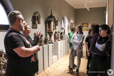 Kolorowe zdjęcie. Po prawej stronie przy ścianie stoi grupa młodzieży. Przygląda się wystawie rzeźb. Po lewej stronie przewodnik muzealny opowiada o wystawie. Obok niego miga tłumaczka.
